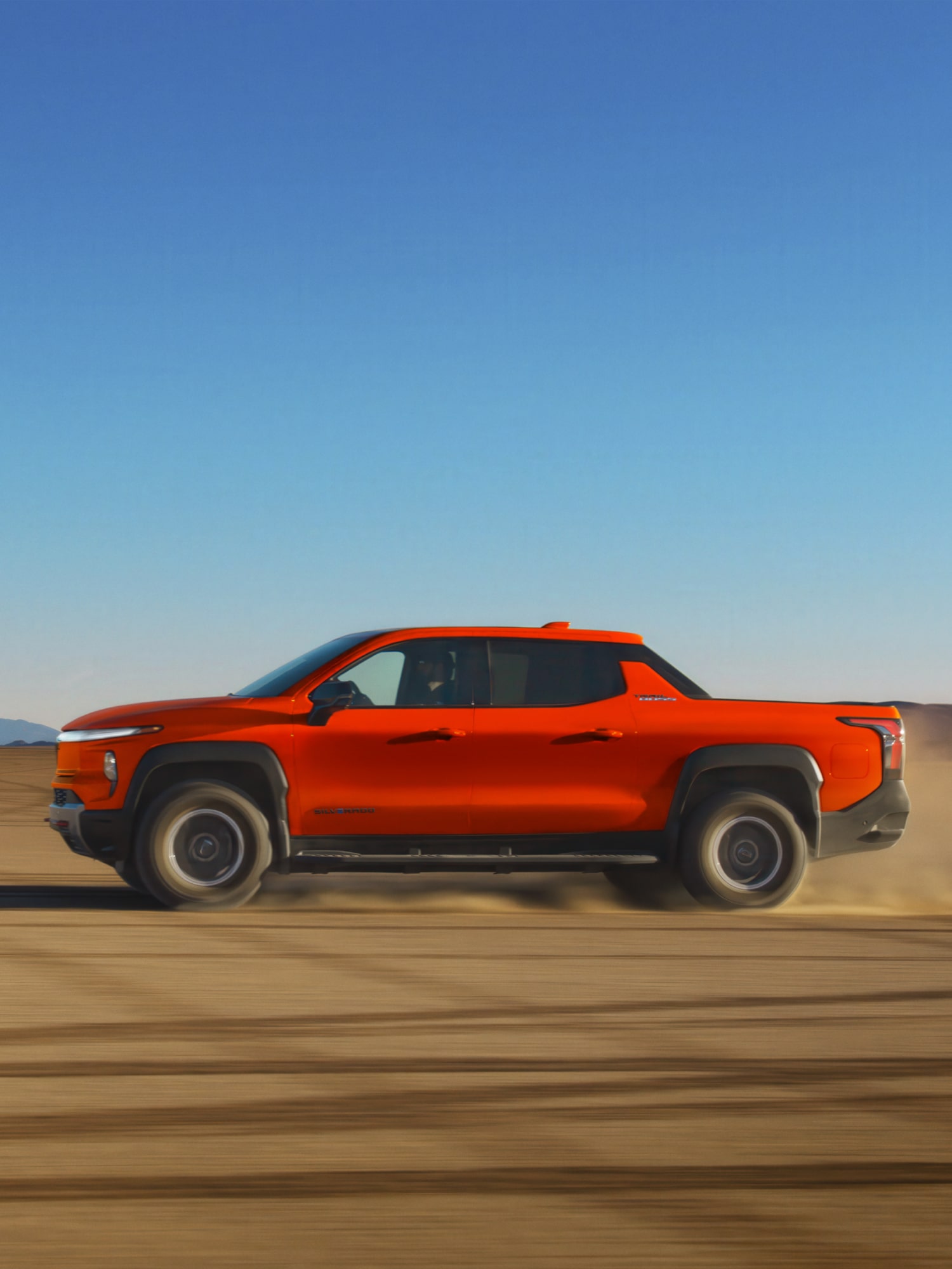 Side View of Orange 2026 Silverado EV Driving on a Dirt Road Highlighting its Off-Road Capabilities and Sleek Design