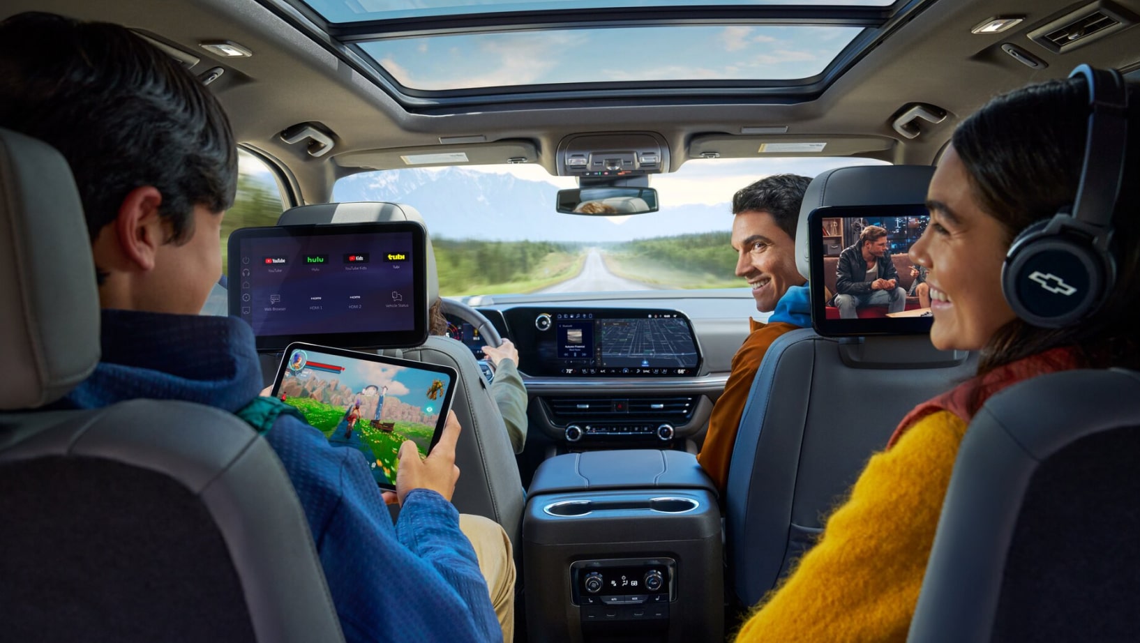 A family of four in a Chevy vehicle with a young boy holding a tablet in the backseat and the other passengers looking at him and smiling.
