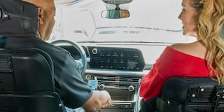 The Interior of a Chevy Traverse SUV Showing Two Front Seats With a View of the Dashboard and Steering Wheel From Behind While Two People Sit Inside.