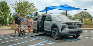 A Silver Chevy Traverse SUV With an Extended Wheelchair Ramp Parked in a Handicap Space Under a Blue Shade Structure While a Group of People Stand Nearby Including One Person With a Wheelchair.