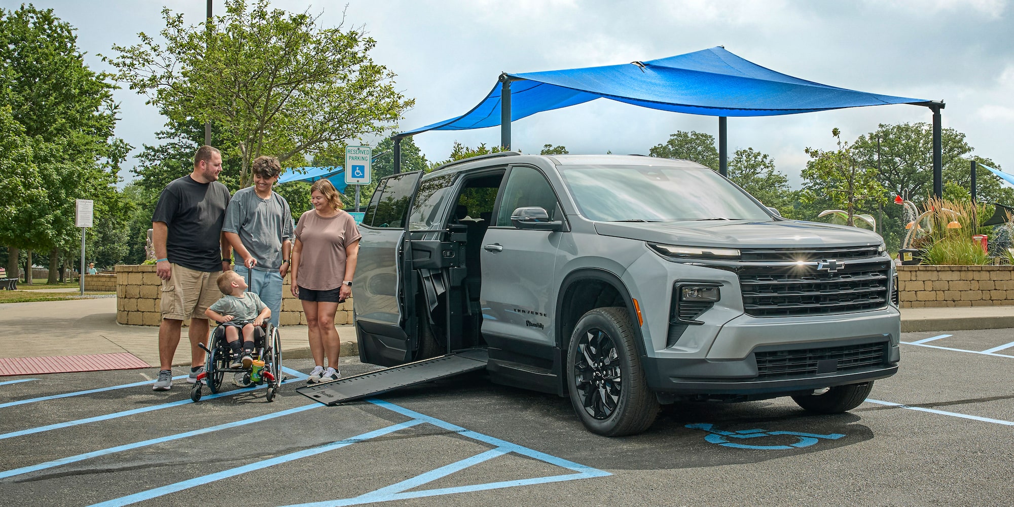 A Silver Chevy Traverse SUV With an Extended Wheelchair Ramp Parked in a Handicap Space Under a Blue Shade Structure While a Group of People Stand Nearby Including One Person With a Wheelchair.