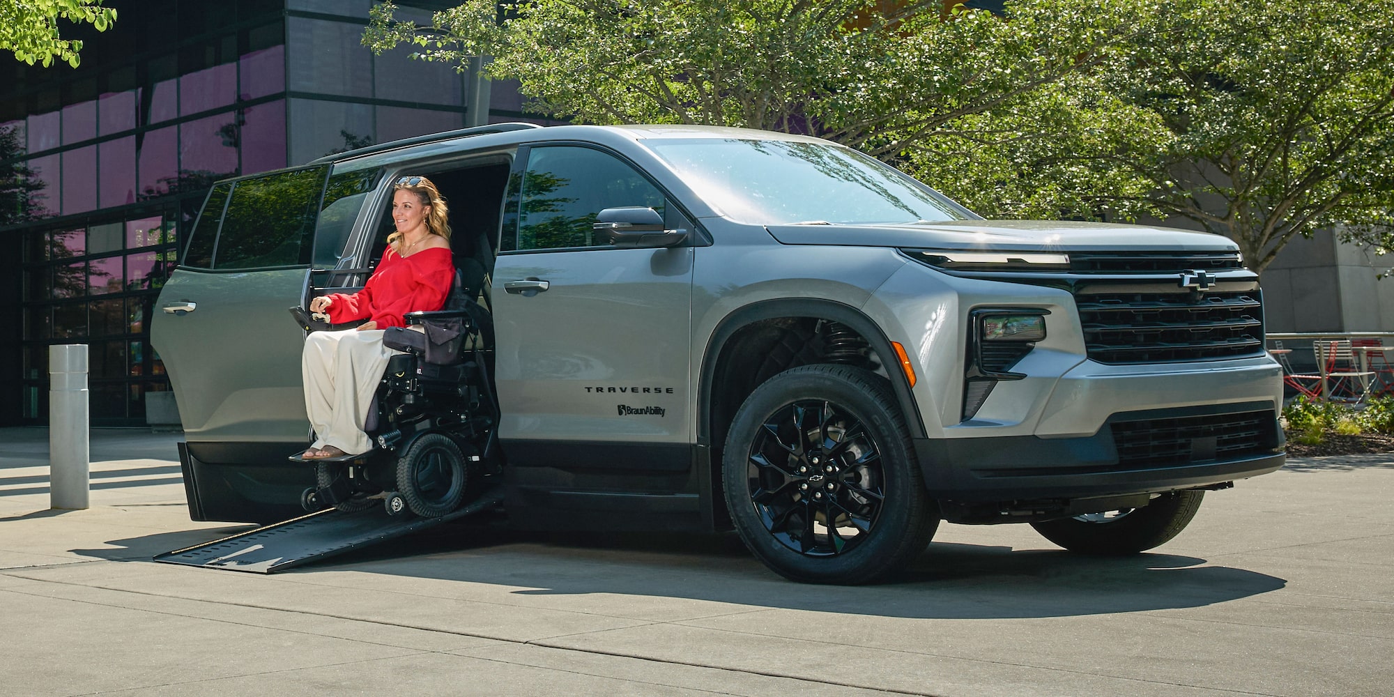 A Silver Chevy Traverse SUV With an Extended Wheelchair Ramp Parked Outside a Modern Building While a Person in a Motorized Wheelchair Wearing a Red Top Uses the Ramp.
