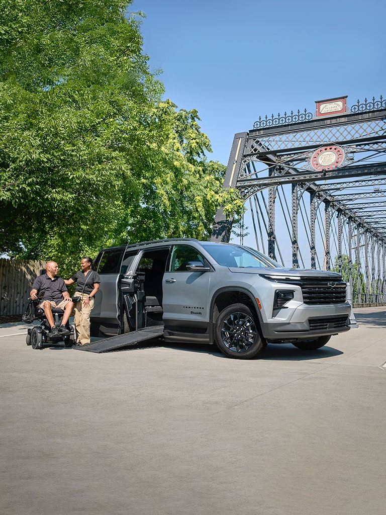 A Silver Chevy Traverse SUV With an Extended Wheelchair Ramp Parked on a Road Beside a Steel Bridge While Two People Converse Near the Ramp.