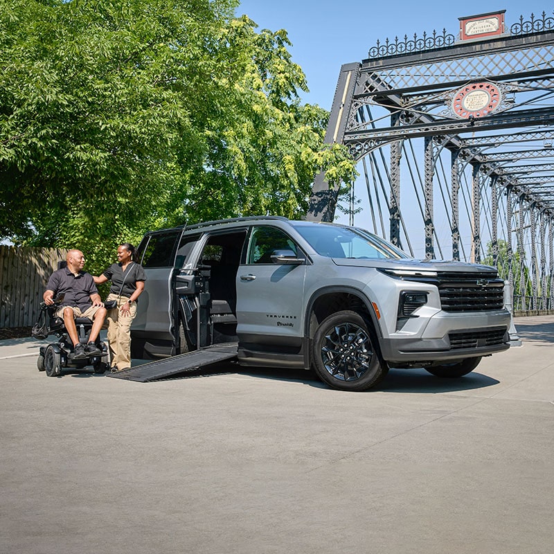 A Silver Chevy Traverse SUV With an Extended Wheelchair Ramp Parked on a Road Beside a Steel Bridge While Two People Converse Near the Ramp.