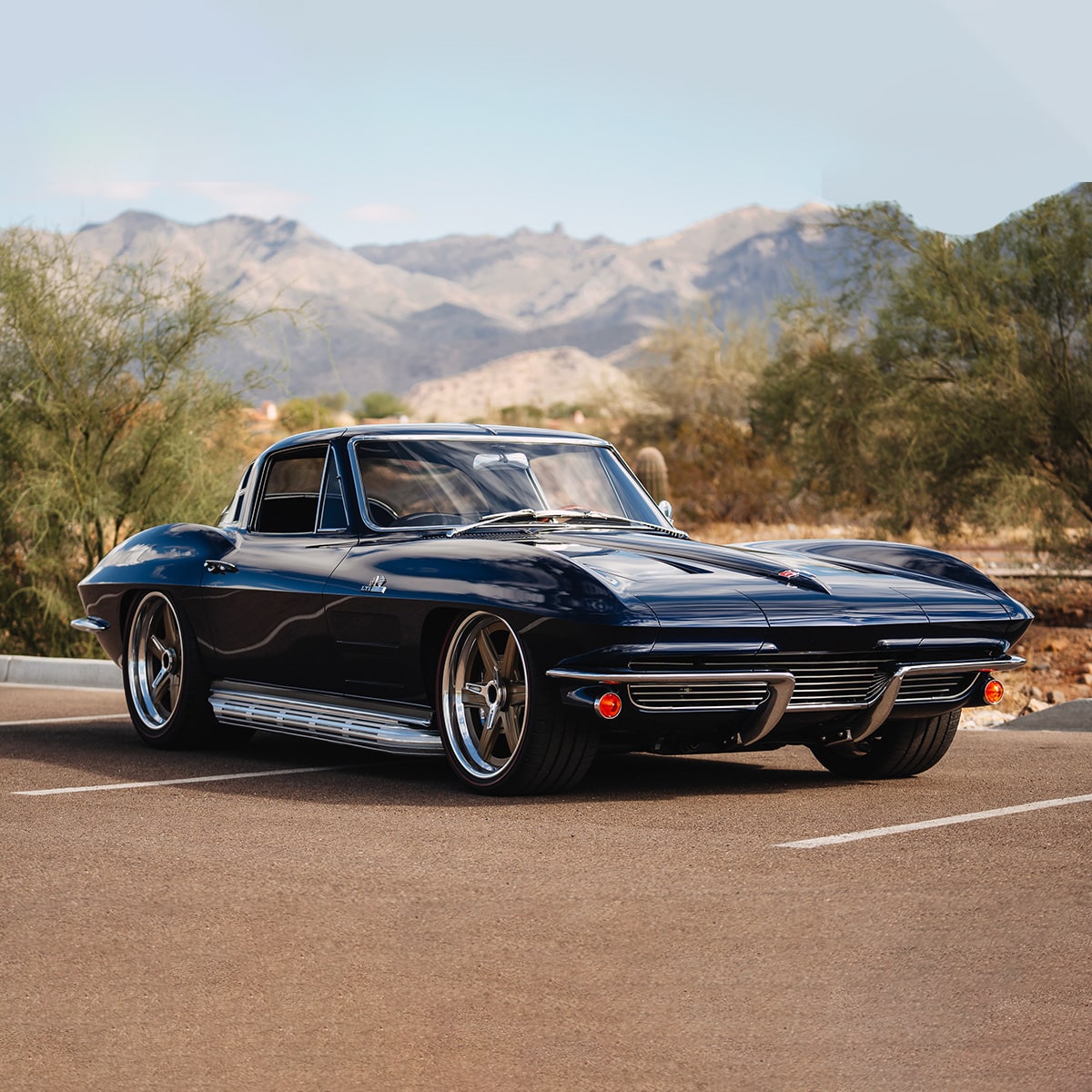 A Dark Blue Classic Chevrolet Corvette Stingray Coupe With Chrome Side Exhaust Pipes Parked on a Paved Road With Mountains and Trees in the Background.