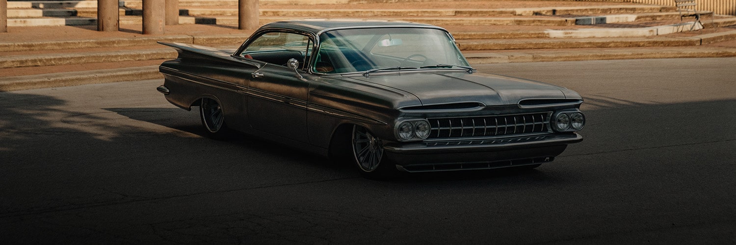 Un Chevrolet Impala clásico de color negro circulando por un camino con un paisaje desértico y montañas de fondo, acompañado por el texto "About Chevrolet Heritage" (Acerca de la herencia de Chevrolet).