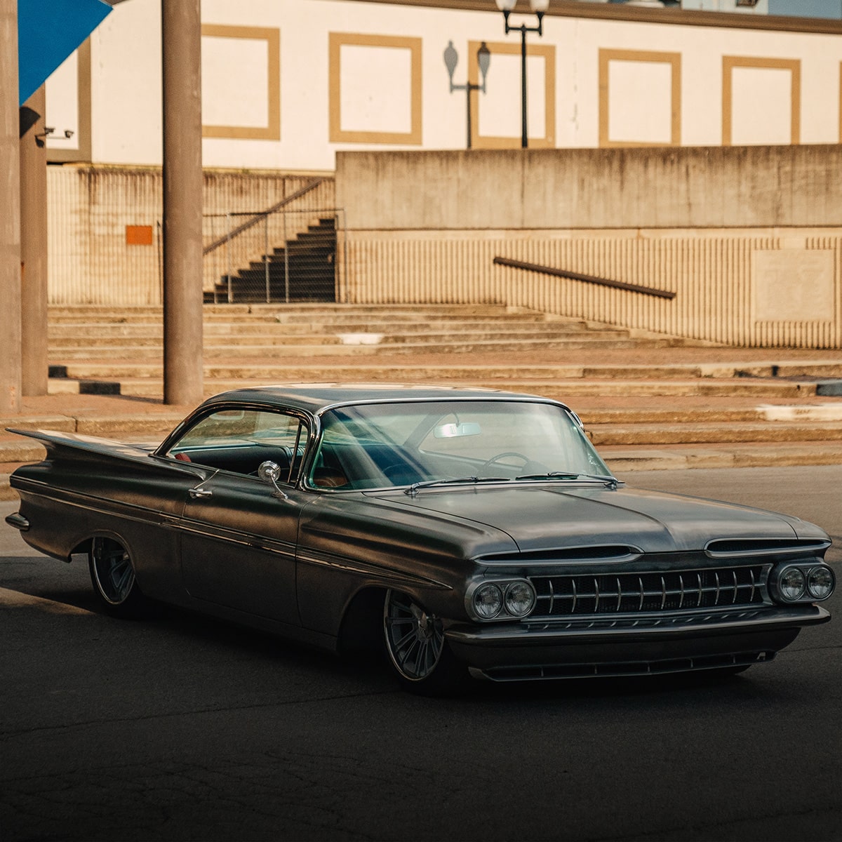 Un Chevrolet Impala clásico negro estacionado en frente de un edificio con altas columnas de piedra y banderas azules.