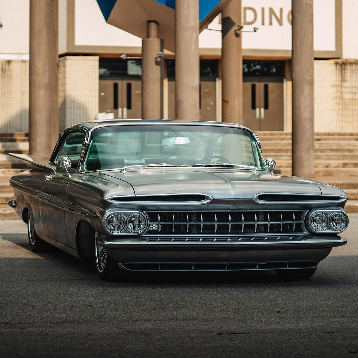 Un Chevrolet Impala clásico negro estacionado en frente de un edificio con altas columnas de piedra y banderas azules.