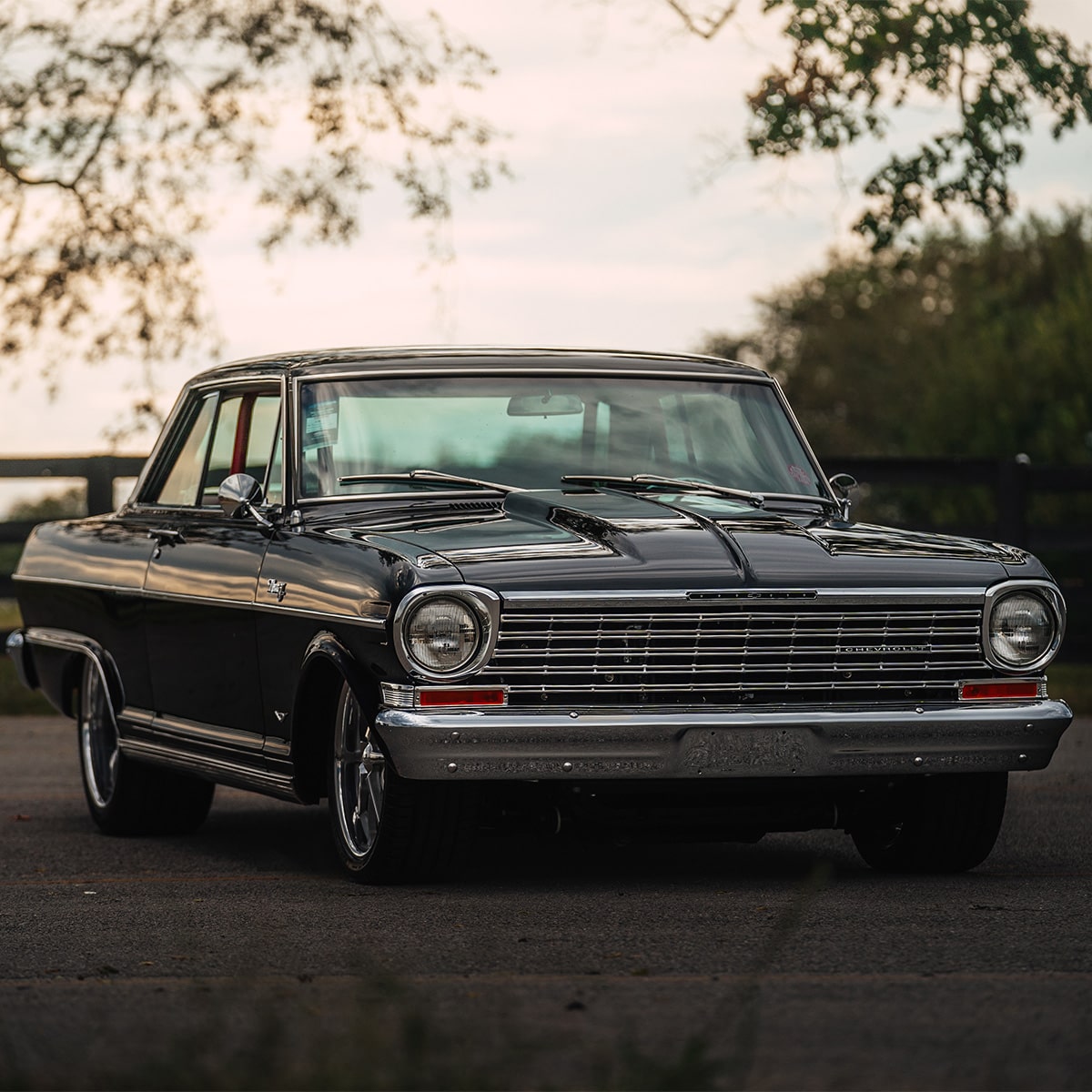 Un Chevrolet Impala negro posicionado al aire libre en un camino con árboles y un atardecer de fondo.