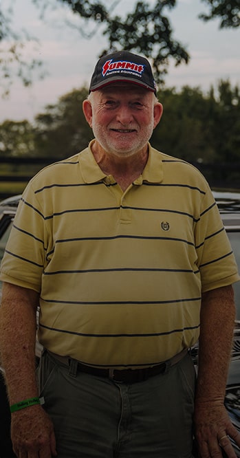 Una persona con una camiseta polo a rayas de color amarillo y verde y una gorra roja parada al aire libre y el texto "My First Car Was a Chevy II and It Got in My Blood" (Mi primer auto fue un Chevy II y lo llevo en la sangre), seguido de "Ronnie Long 1966 Chevy II Nova" (Chevy II Nova de Ronnie Long).