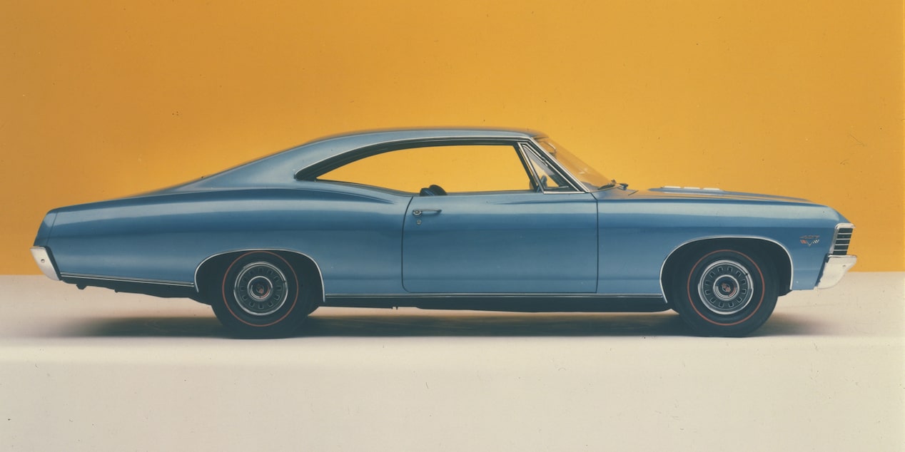 Chevrolet Impala azul estacionado al aire libre en una entrada vehicular, con arbustos verdes al fondo.