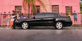 Chevrolet Impala negro estacionado frente a un edificio rosa intenso, con palmeras y una cerca metálica.