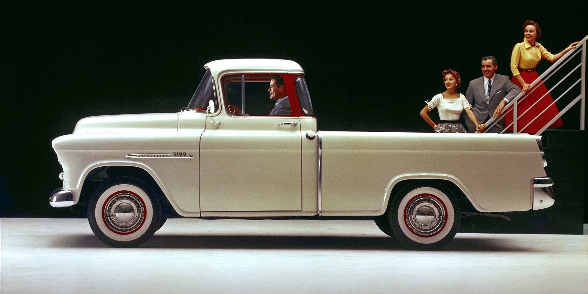 A Classic Cream Chevrolet Silverado 1500 Pickup Truck Parked on a Road With a Person Standing Beside It and Trees in the Background.
