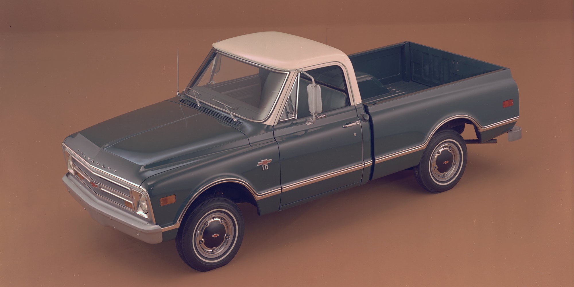 A Teal Chevrolet Silverado 1500 Pickup Truck Parked on a Road With a White Roof and Chrome Accents Against a Brown Landscape.