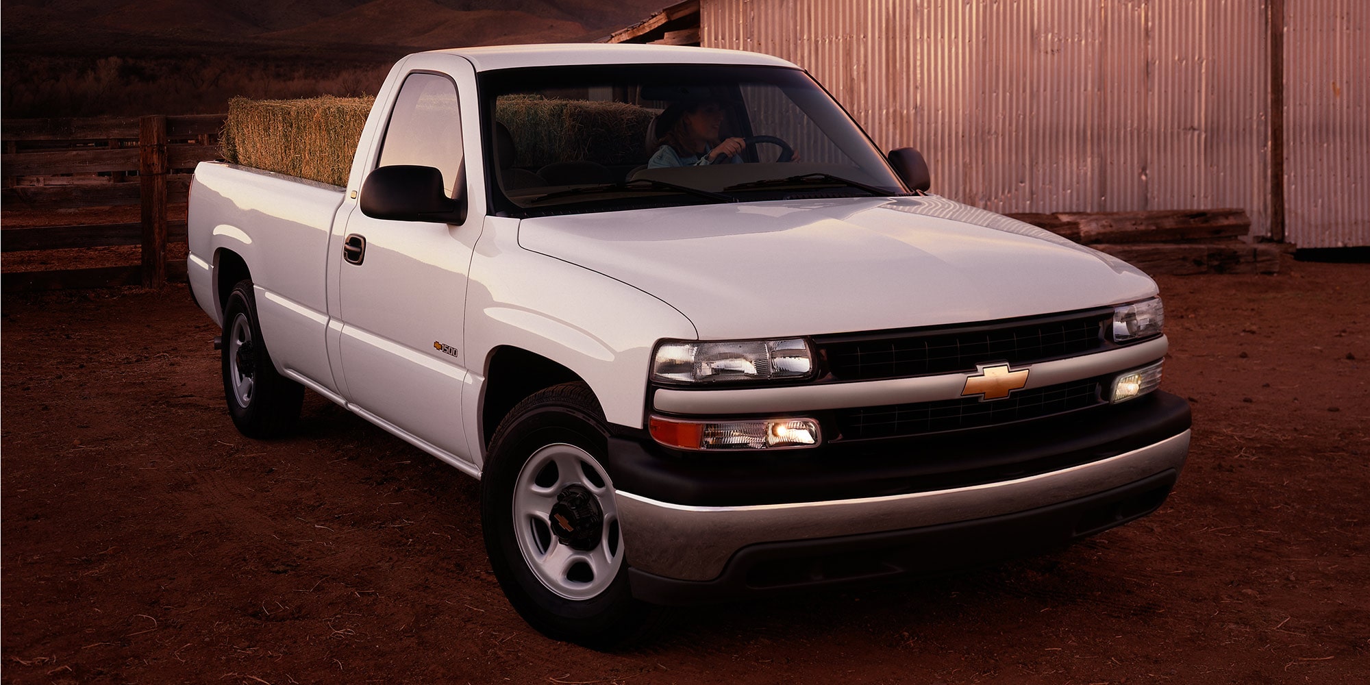 A White Chevrolet Silverado 1500 Pickup Truck Parked on a Dirt Surface With a Brick Wall and Warm Lighting in the Background.