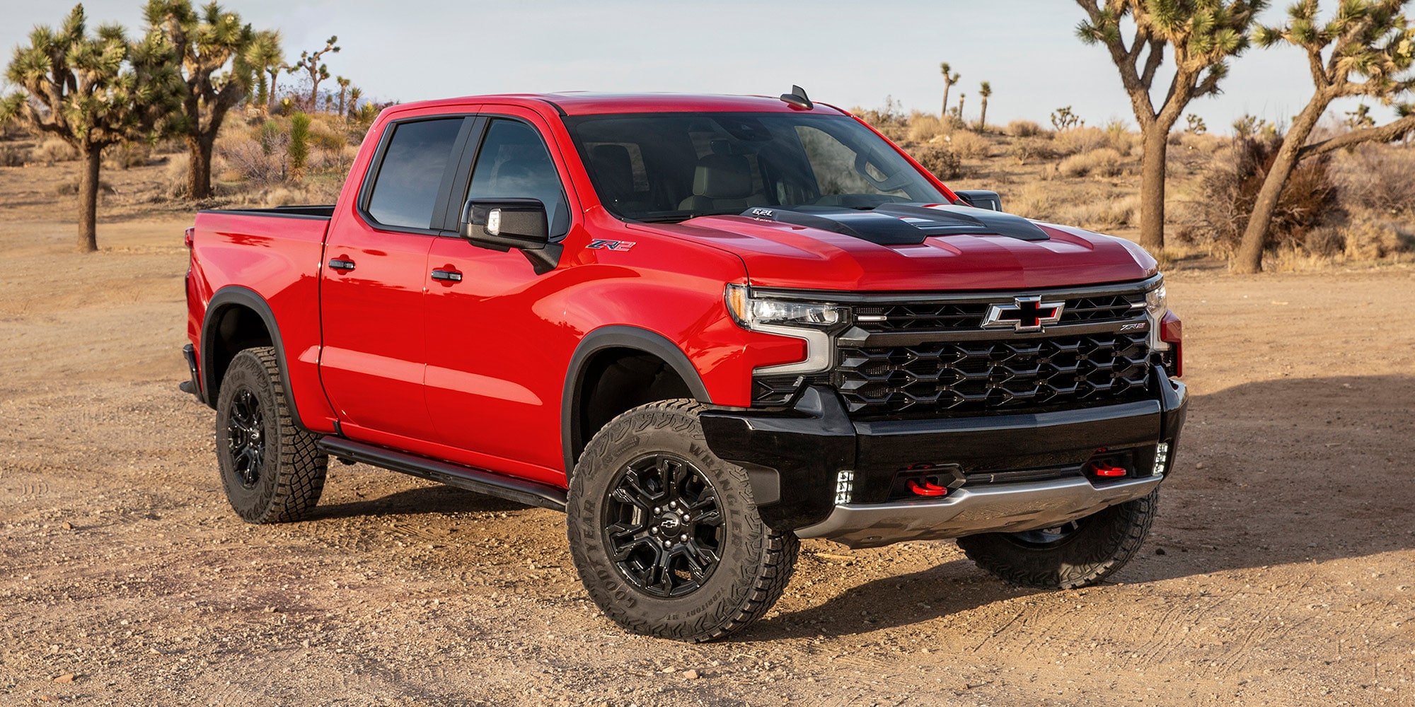 A Red Chevrolet Silverado 1500 ZR2 Parked on a Dirt Surface With Desert Vegetation and Trees in the Background.