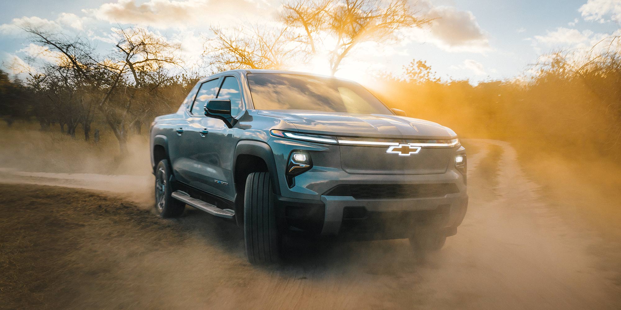 A Silver Chevrolet Silverado EV Driving Through a Dusty Terrain With Sunlight Breaking Through Clouds in the Background.