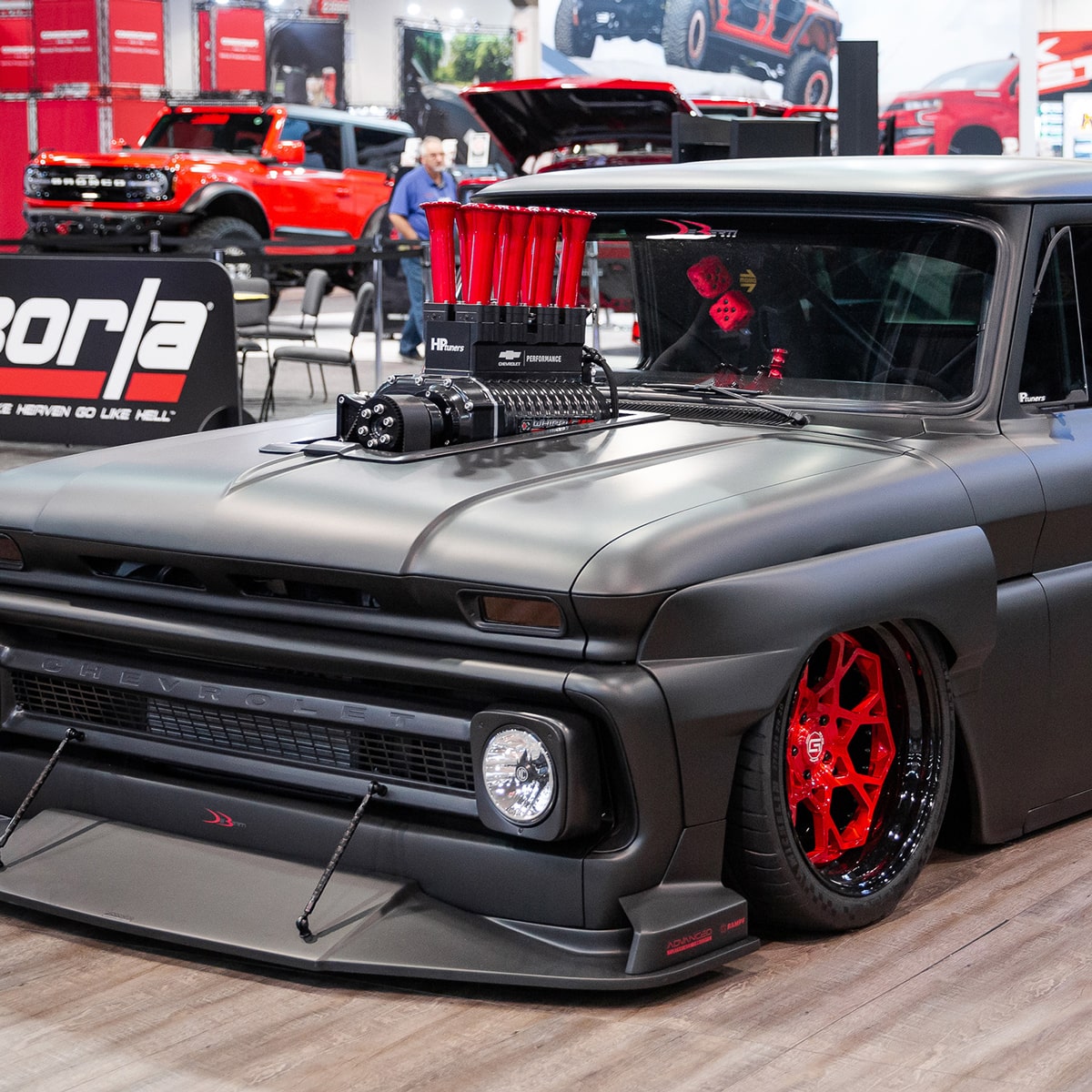 A Matte Black Chevrolet Black C10 Slayer with a Prominent Supercharger and Red Velocity Stacks on the Hood, Wide Body Kit, and Red Deep-Dish Wheels Displayed Indoors at an Automotive Event.