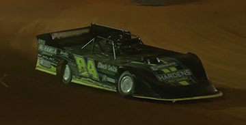 A Black Dirt Track Race Car with the Number 24 on its Side Speeds Around a Curve on a Dirt Track at Night, Kicking up Dust. A Play Button Overlay Suggests this is a Video Still.