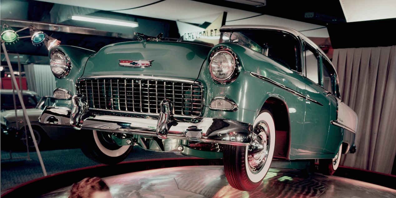 A Green 1955 Chevy Bel Air with the Front Side Held in the Air in a Display Room with a Woman in a Red Dress Sitting in Front of It