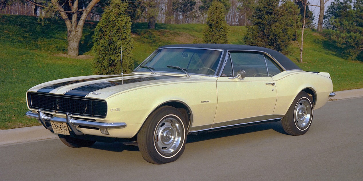 A 1967 Chevrolet Camaro SS with Vertical Black Stripes on the Hood with Grass and Trees in the Background