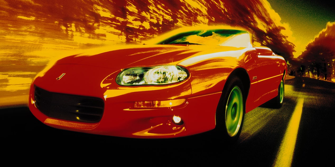 A Red 1998 Chevy Camaro Z28 Driving Down the Road with Red Trees Stylishly Blurring as It Zips by Them