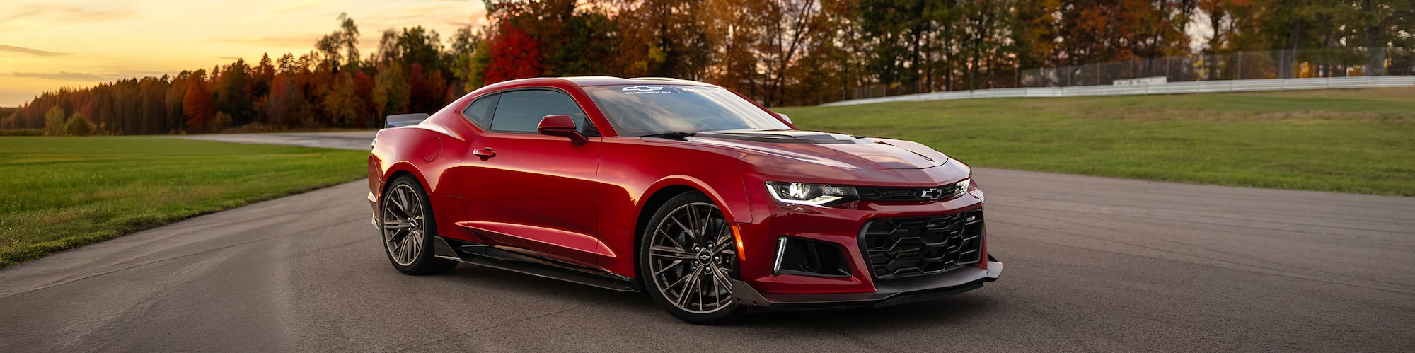 Red Chevrolet Camaro Sports Car Parked on a Racetrack with Autumn Trees and a Sunset Sky in the Back