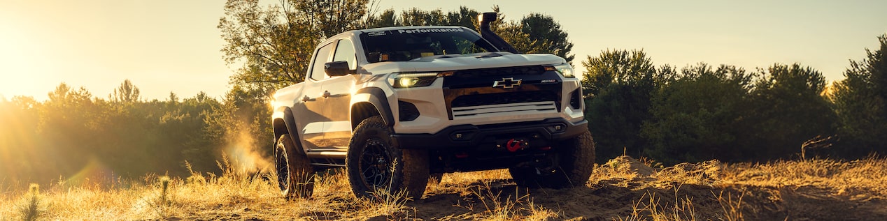 Front Three Quarters View of a White Chevrolet Colorado on a Dirt Path Near a Forest with the Sun Setting in the Horizon