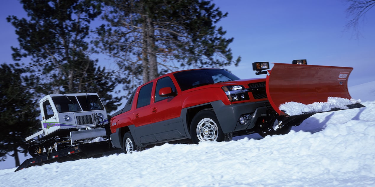 A Red 2002 Chevy Avalanche Pickup Truck Equipped with a Snow Plow Driving Ahead a Snowmobile