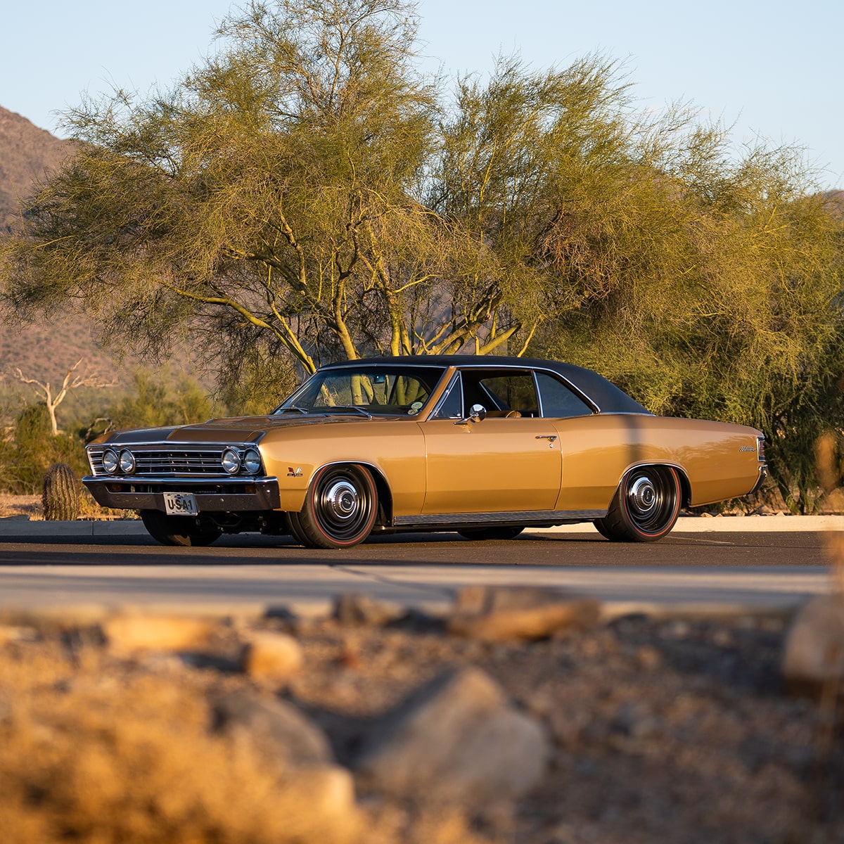 A Gold 1967 Chevy Malibu Parked on a Scenic Road with Mountains and Trees in the Background