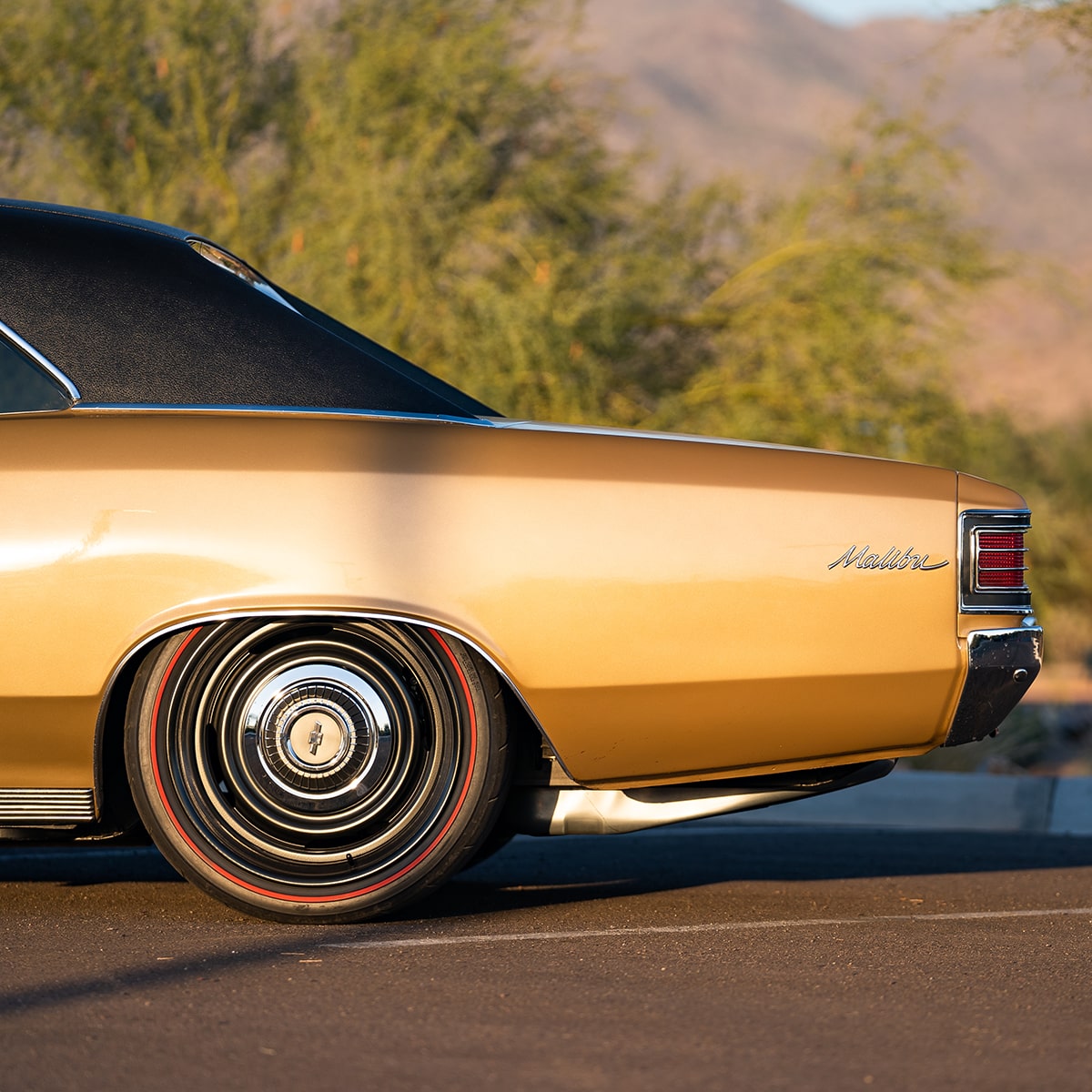 Rear Left Half Side View of a Gold 1967 Chevy Malibu in a Parking Lot with Trees and Mountains in the Distance