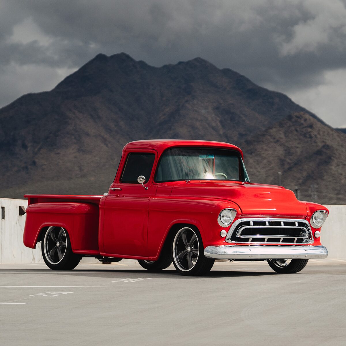 Distant View of a Bright Red 1957 3100 Chevy Pickup Truck in a Parking Lot with Mountains and Grey Clouds in the Distance