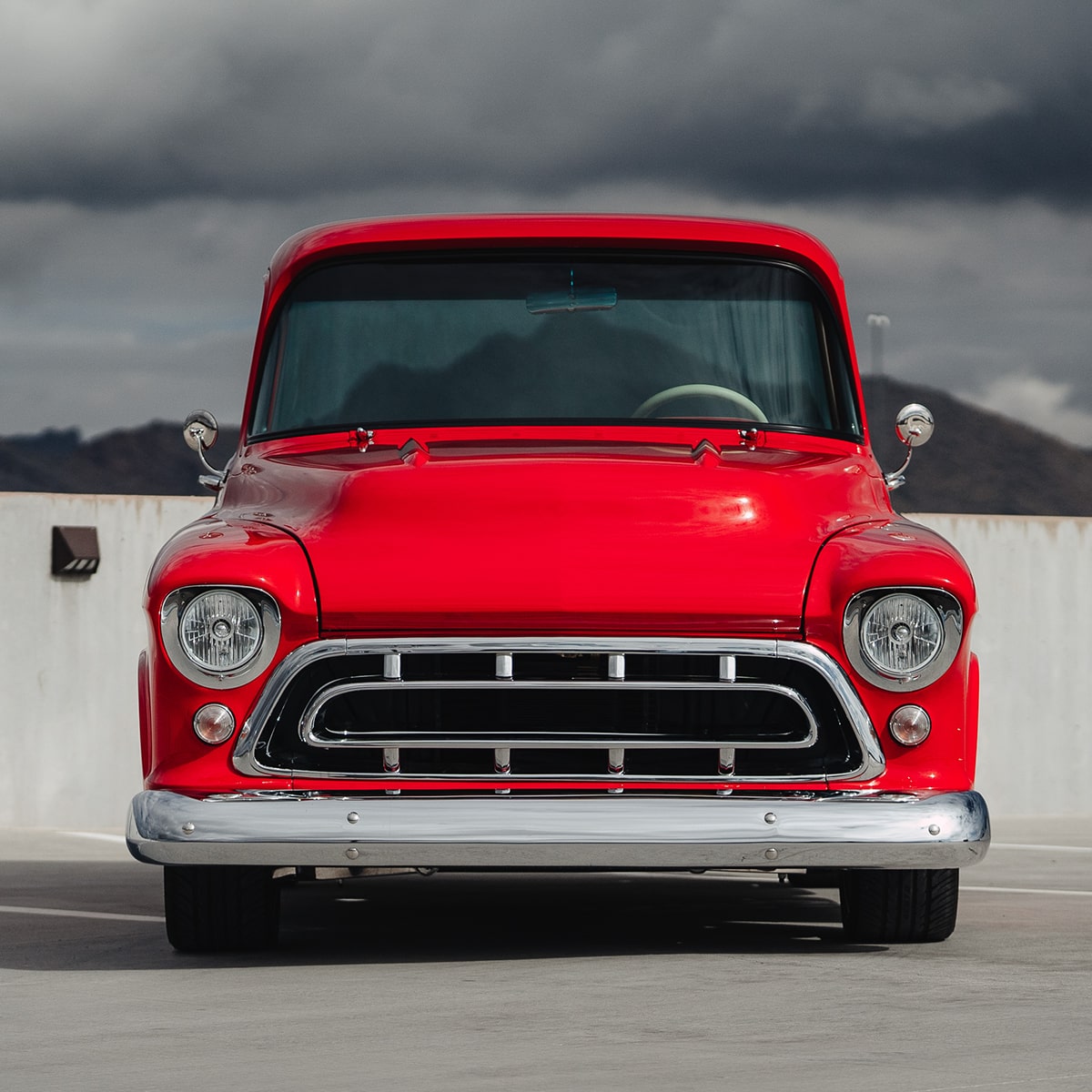 Front View of a Bright Red 1957 Chevy 3100 Pickup Truck in a Parking Lot with Mountains and Grey Clouds in the Distance