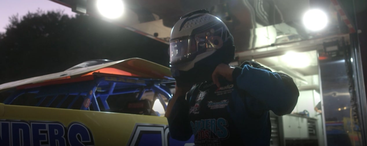 A race car driver wearing a helmet and blue racing suit prepares for a race in a well-lit garage. The text "Chevrolet Performance Parts" appears in the lower-left corner, with a play button overlay indicating this is a video still.