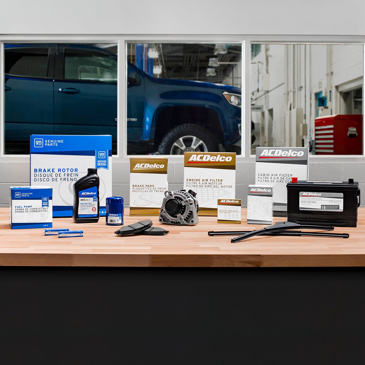 A Table with a Variety of Vehicle-Related Products such as Motor Oil and a Car Battery Beneath Three Windows Looking into an Auto Shop with a Blue GM SUV