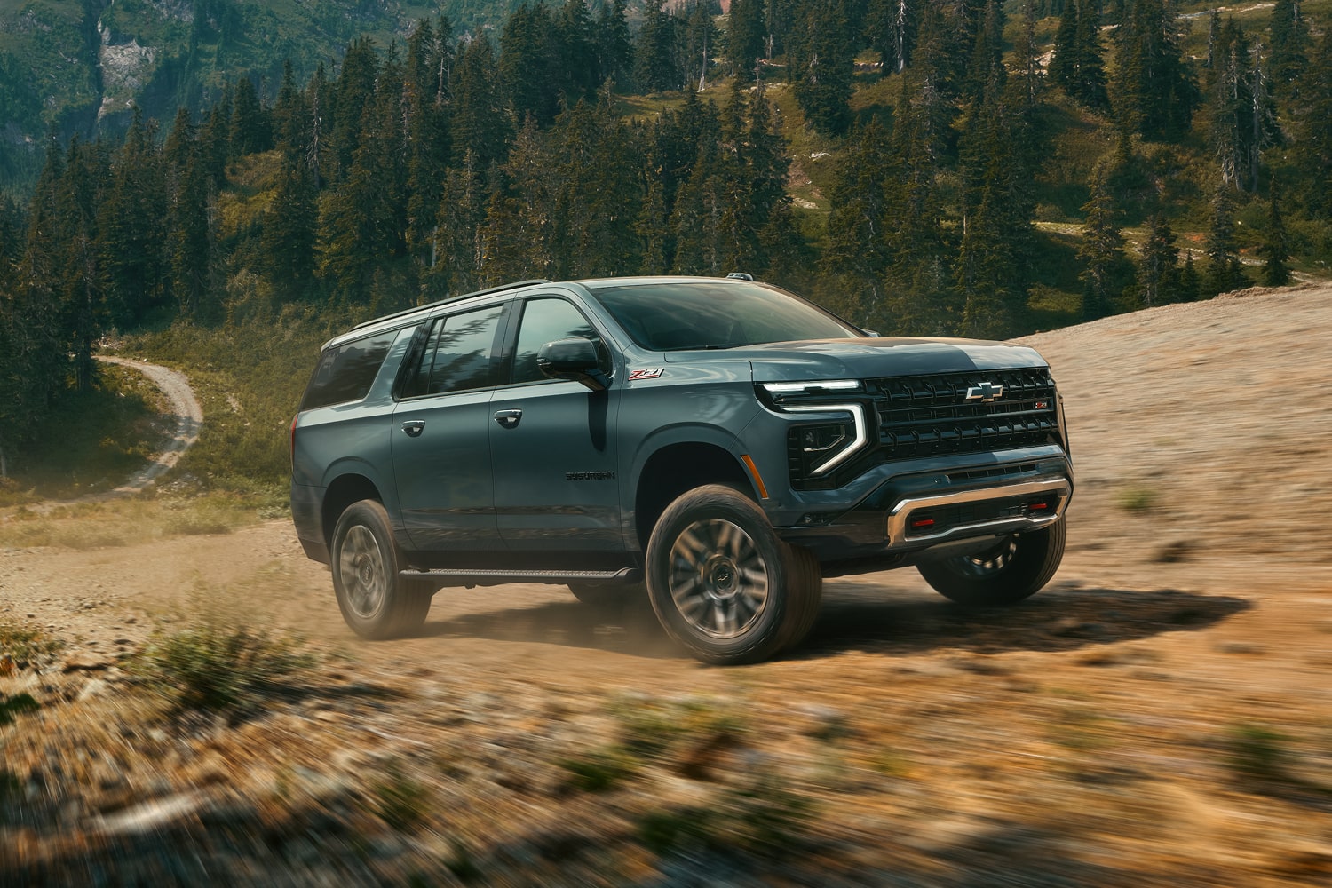 A Chevrolet Suburban Kicking Up Dust While Driving Off-Road Through a Rocky Trail With a Forested Mountain Landscape in the Background.