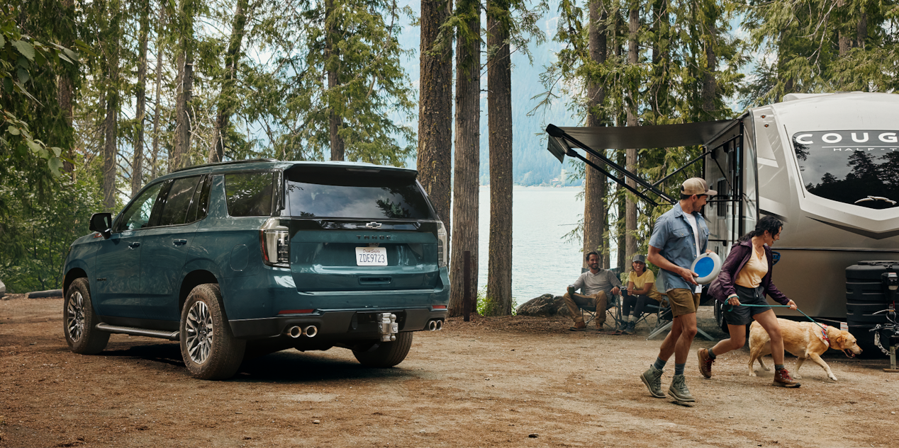 Three-quarters Rear View of the 2026 Chevy Tahoe Parked in the Woods Next to a Family Playing by Trailer