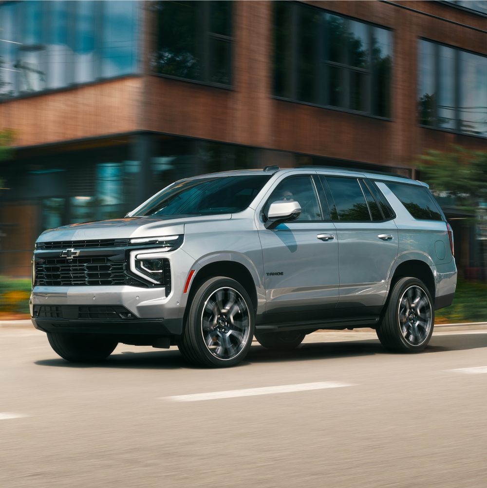 Profile View of the 2026 Chevy Tahoe SUV Driving Past a Modern Apartment Building
