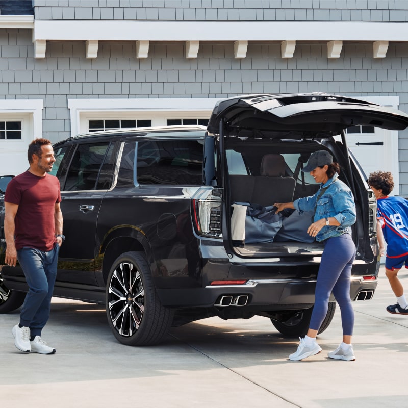 Suburban High Country shown in Dark Ash Metallic parked in a drive way with a family surrounding an open trunk.