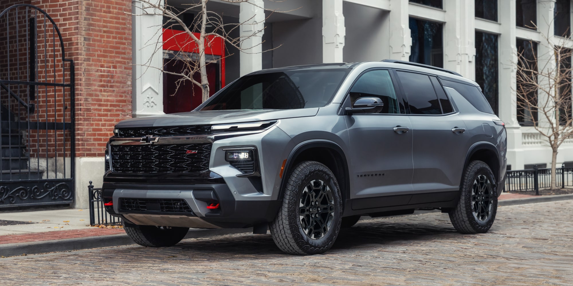 A 2027 Chevy Traverse Parked on a City Street Showcasing a Bold Front Grille, Black Alloy Wheels, and Rugged SUV Styling From a Front Three-Quarter Angle.
