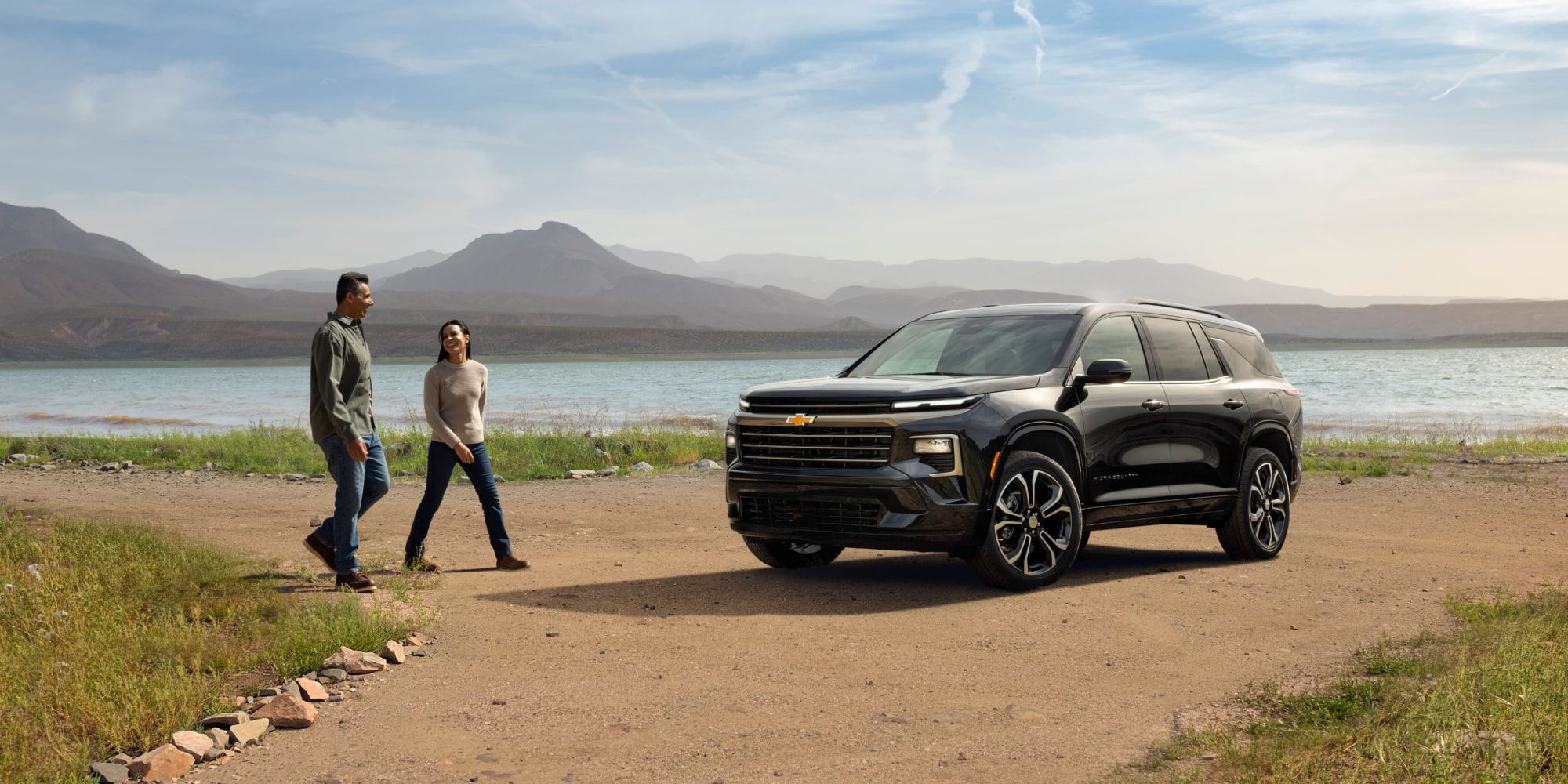 A Couple Talking and Walking Towards a 2027 Chevy Traverse Parked by a Lake Surrounded by the Mountains
