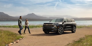A Couple Talking and Walking Towards a 2027 Chevy Traverse Parked by a Lake Surrounded by the Mountains