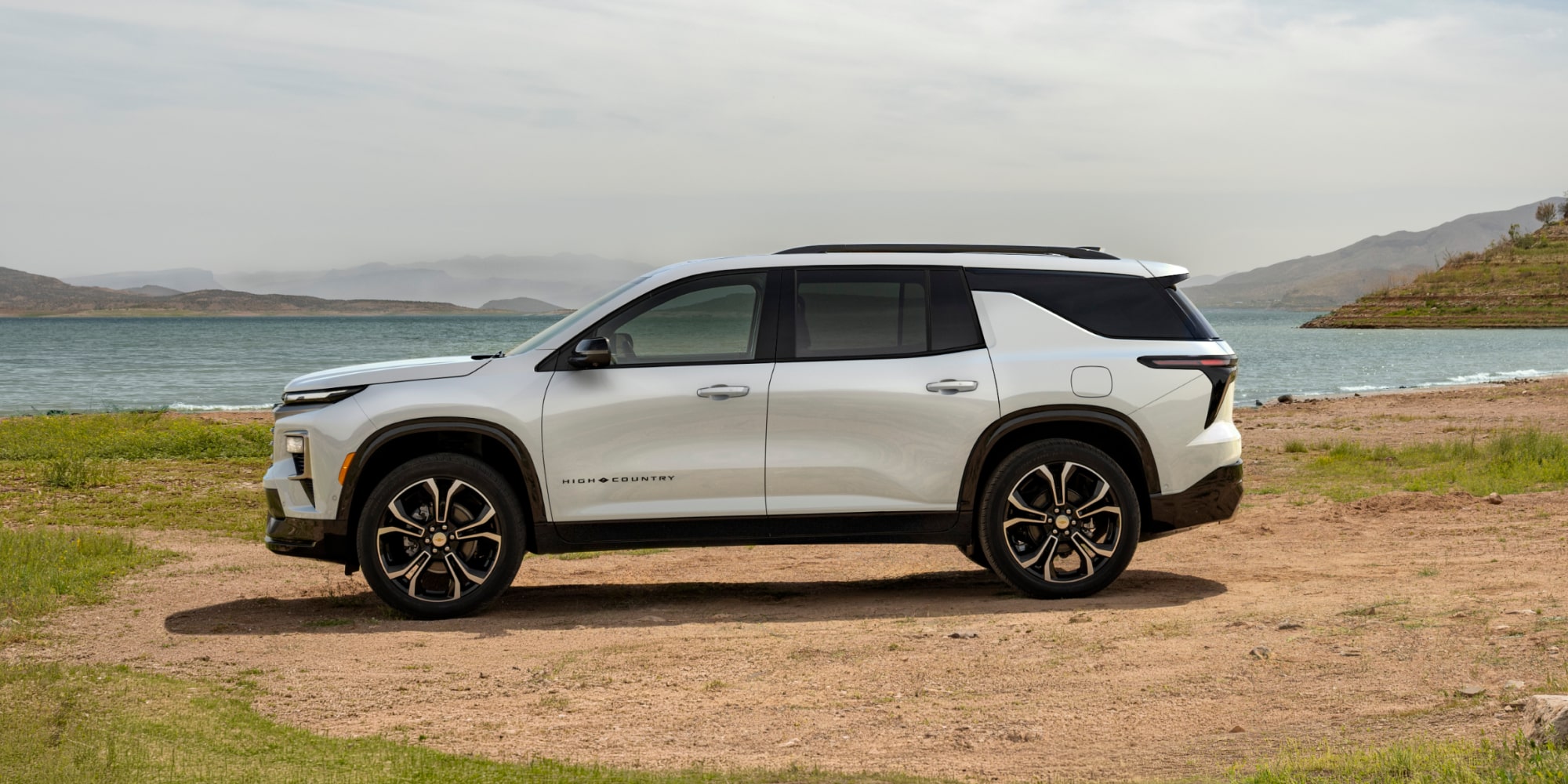 A Profile View of a White 2027 Chevy Traverse Parked on a Dirt Patch Next to a Lake