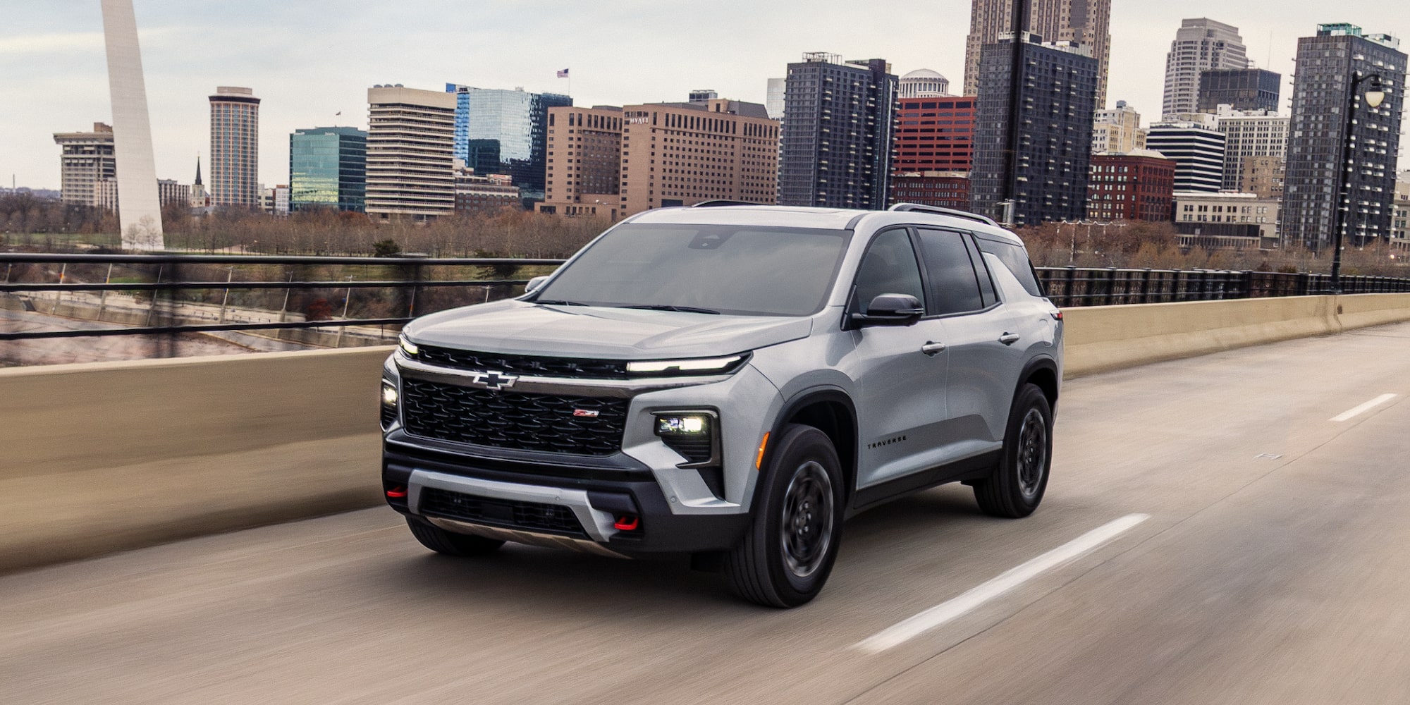 A 2027 Chevy Traverse Driving on an Urban Highway With a Modern City Skyline in the Background.