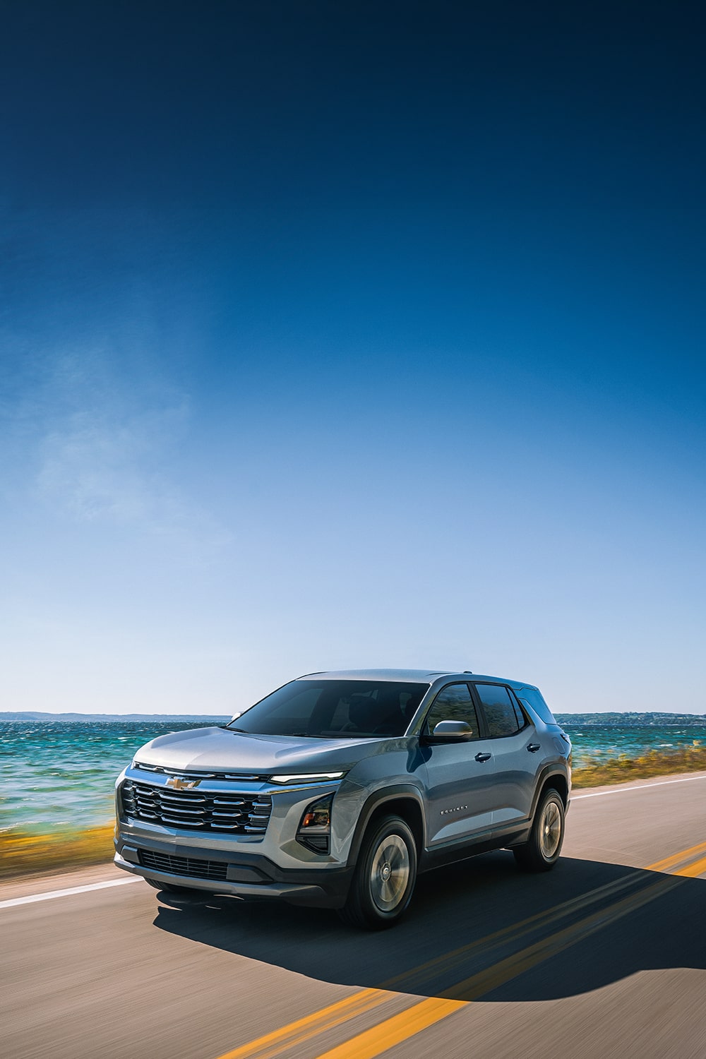 A Green Chevrolet Equinox Driving Down a Sunlit Rural Road With Blurred Trees and Hills in the Background.