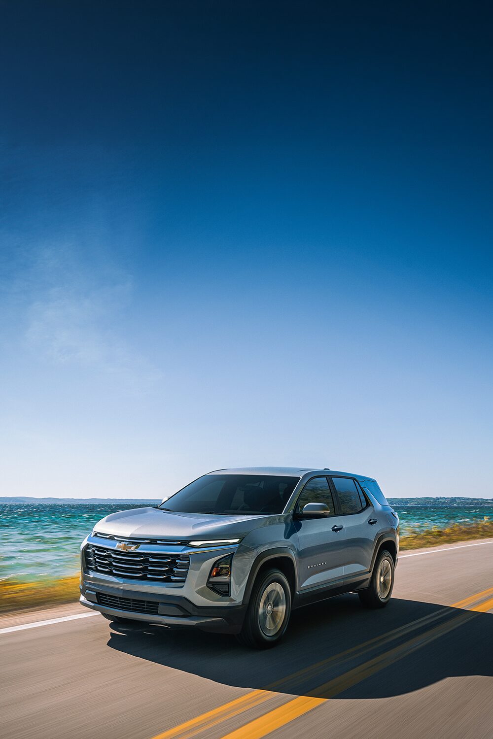 A Green Chevrolet Equinox Driving Down a Sunlit Rural Road With Blurred Trees and Hills in the Background.