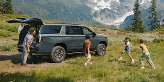 A Chevrolet Suburban Parked Beside a Mountain Meadow as Children Run and Play Near Tall Evergreen Trees.