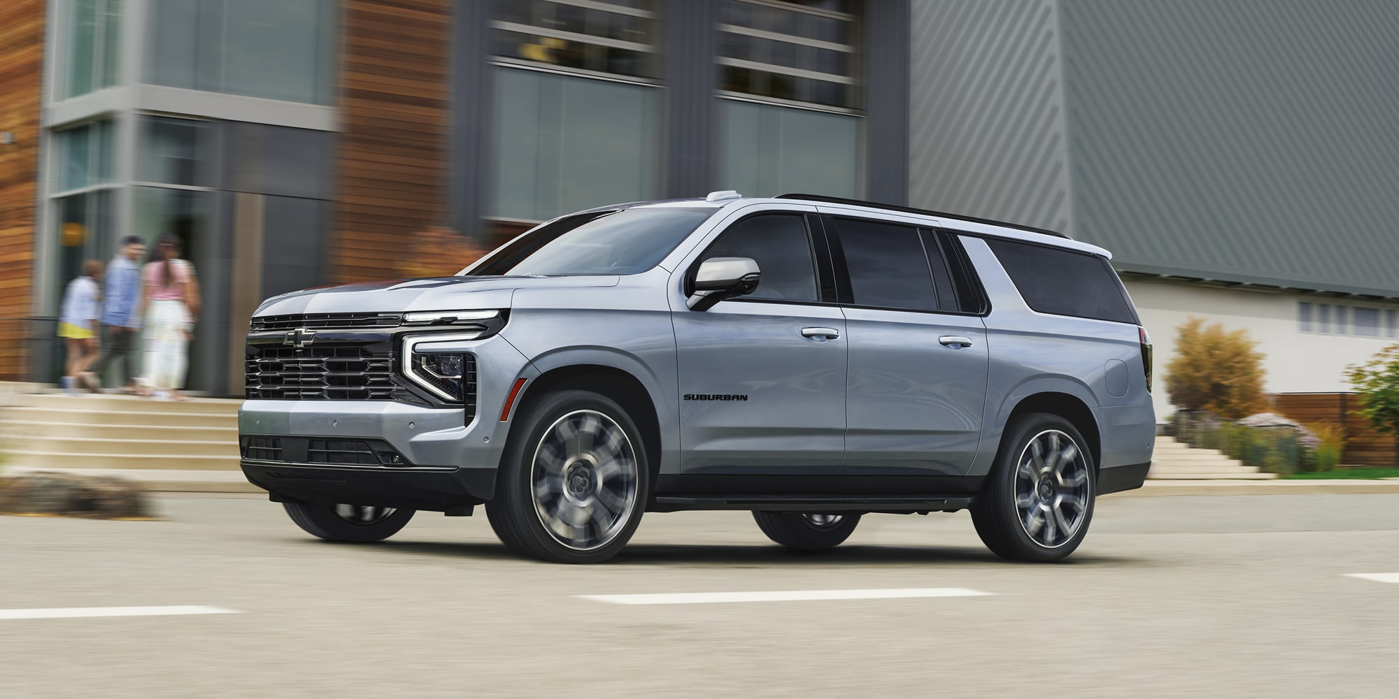 A Silver Chevrolet Suburban Driving Through an Urban Area Past Modern Buildings and Storefronts.