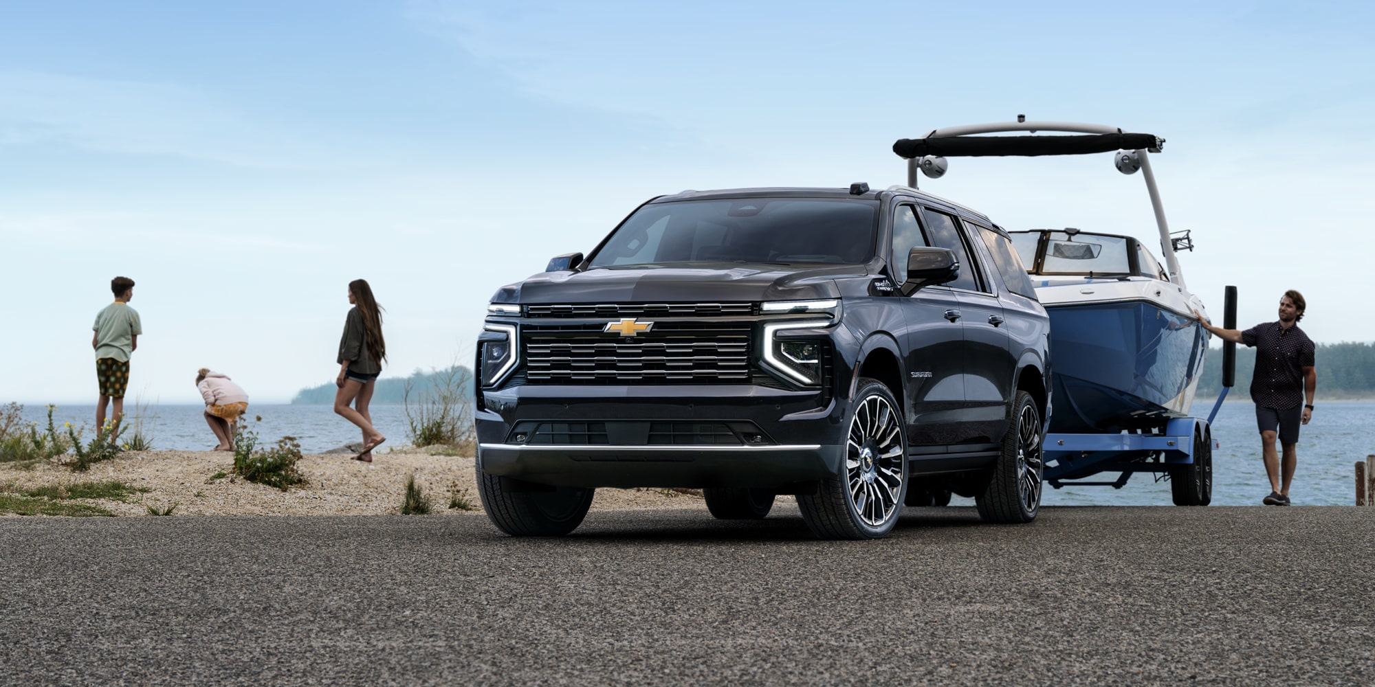 A Black Chevrolet Suburban Parked on a Rocky Lakeside Shore While Towing a Blue Boat Beside People Standing Near the Water.