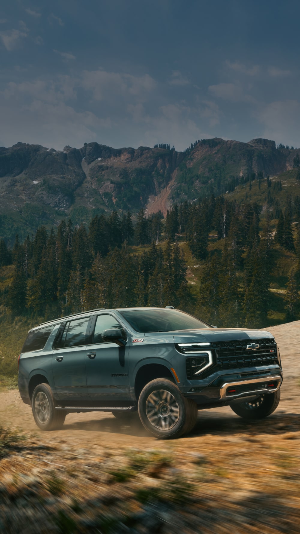 A Black Chevrolet Suburban Driving Along a Rugged Mountain Path Surrounded by Golden Grass, Evergreen Forests, and Rolling Hills in Warm Evening Light.